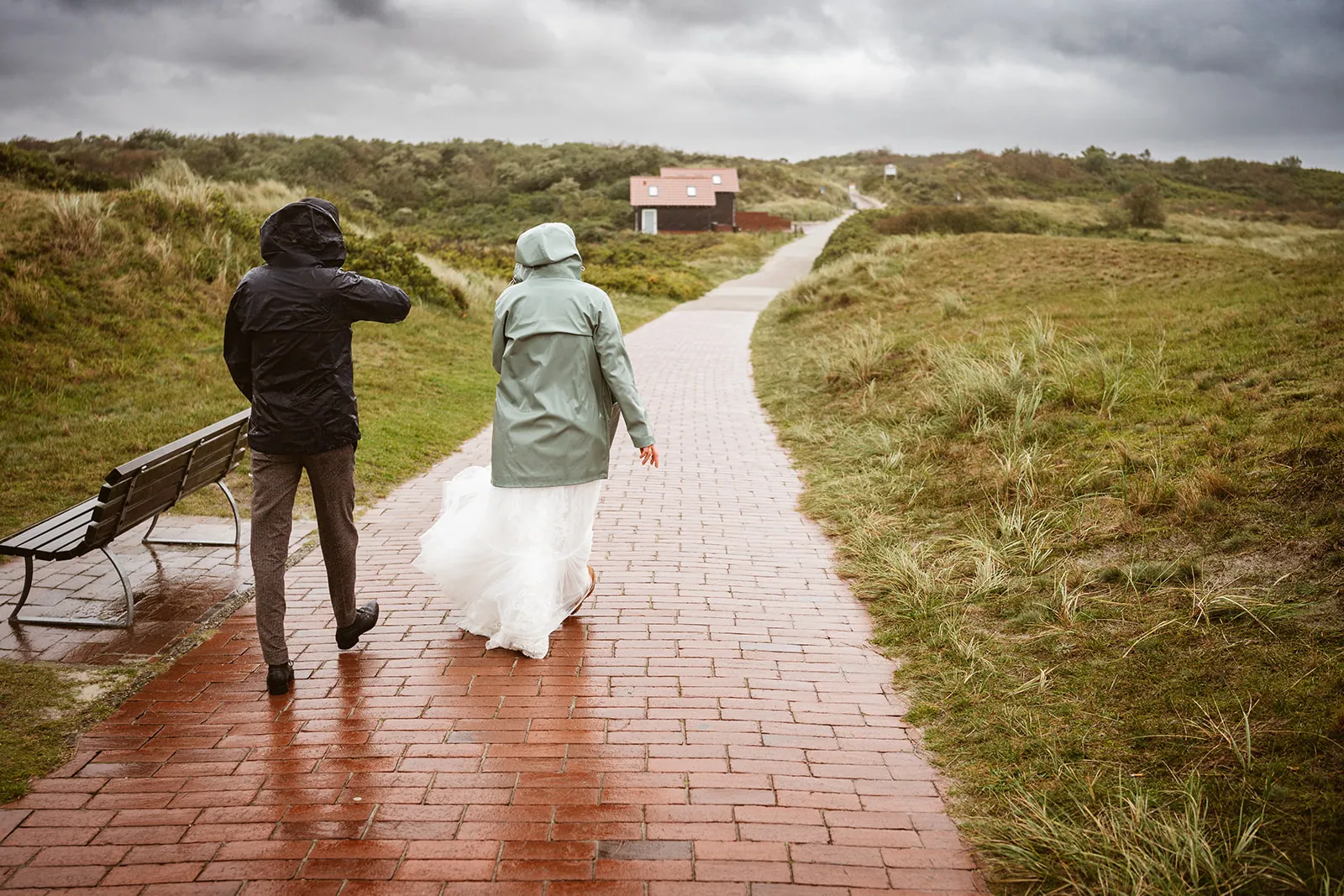 Mel und Christoph gehen in Regenjacken über den nassen Dünenweg auf Langeoog