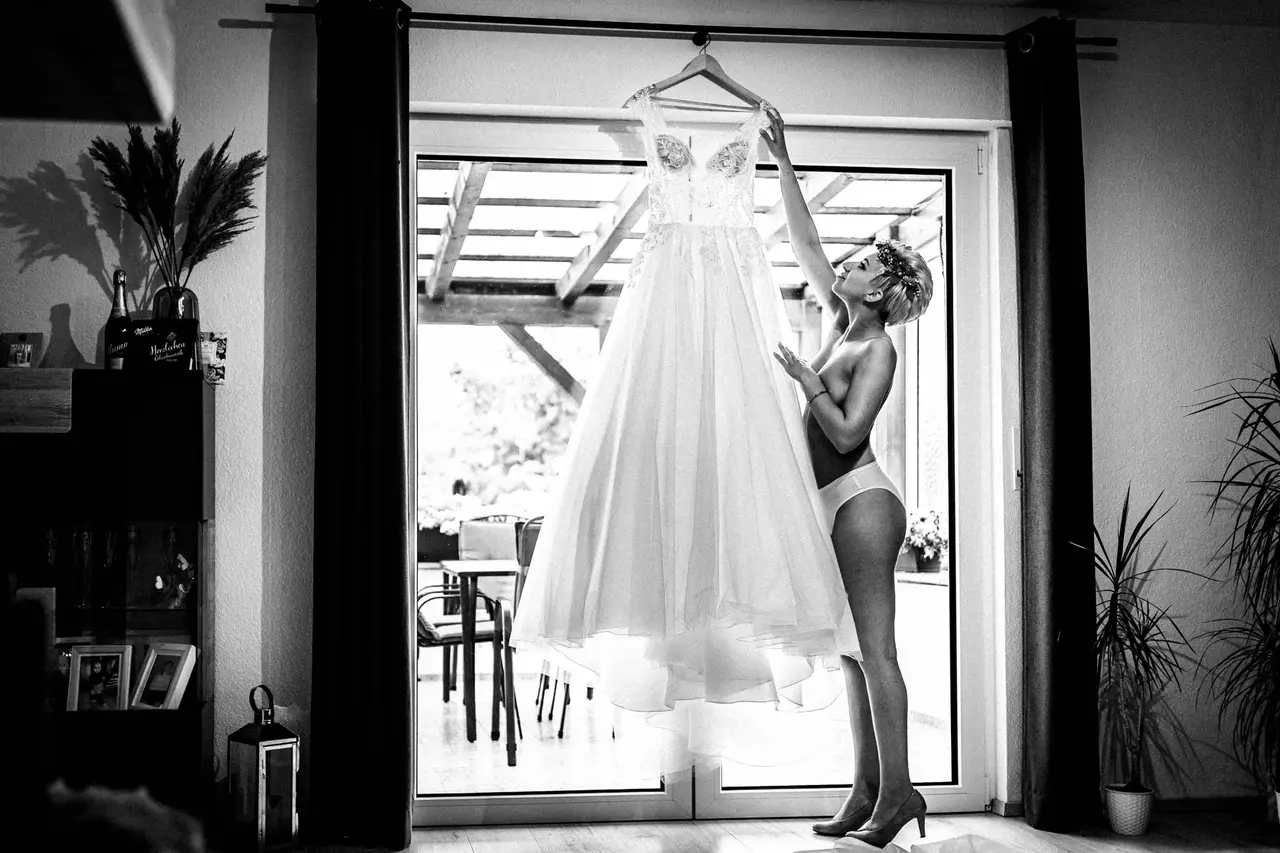 Braut hebt beim Getting Ready ihr Kleid vor dem hellen Fenster an