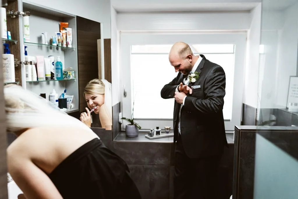 Braut putzt im Bad die Zähne, während der Bräutigam sich für die Hochzeit fertig macht